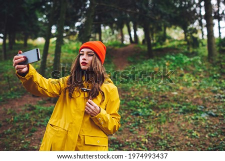 Low angle of young female in yellow raincoat and red hat standing in fall forest and taking selfie using smartphone