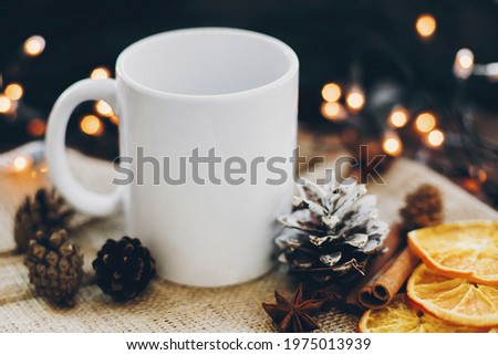 White empty cup and pine cones, anise, cinnamon, dried oranges on background of christmas lights. Winter time for hot cocoa and tea. Christmas holidays