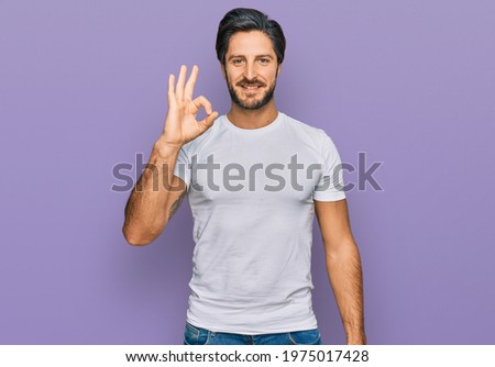 Young hispanic man wearing casual white t shirt smiling positive doing ok sign with hand and fingers. successful expression. 