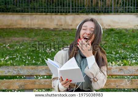Young woman in white electronic headphones laughing and listening online audiobook in nature. Enjoying music or comic humoristic podcast. Storytelling. Leisure or spare time concept
