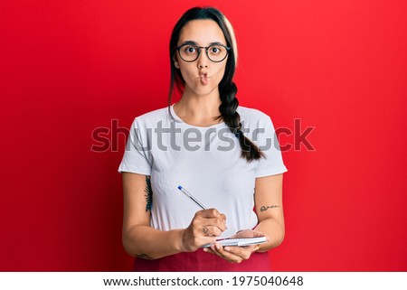 Young hispanic woman wearing waitress apron taking order making fish face with mouth and squinting eyes, crazy and comical. 