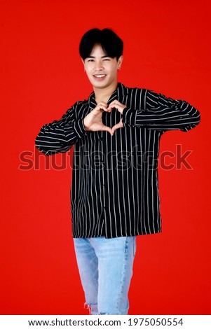 Portrait asian man about 20-25 years old standing while his hands make to heart symbol with looking at camera feel happy  on face. In studio with red background with concept valentine of love