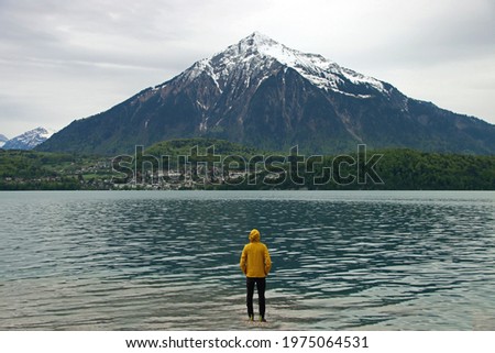 Man with yellow rain jacket in front of huge snow-covered mountain in Switzerland. Mount Niesen with green trees and water of lake Thun. Royalty-Free Stock Photo #1975064531