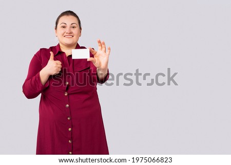 Smiling cheerful young woman 30s in bordo casual dress standing hold empty blank mock up credit bank card showing thumb up looking camera isolated on gray background, studio. High quality photo