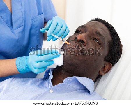 Closeup face of adult African American male client receiving injections during lip enhancement procedure, professional cosmetologist hands in rubber gloves holding syringe..