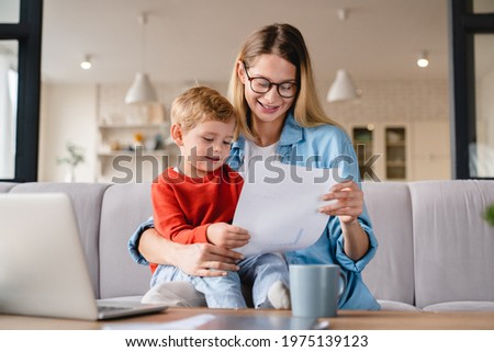 Little small toddler kid child son helping her busy young mother with freelance remote work holding papers, documents and domestic bills at home on lockdown during maternity leave Royalty-Free Stock Photo #1975139123