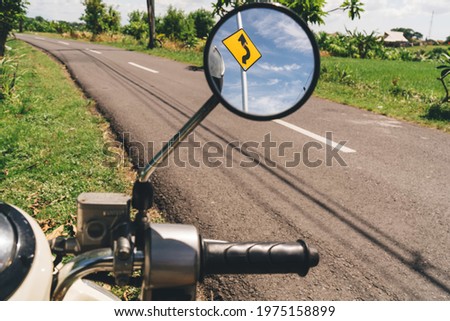 Road curve traffic sign reflecting in round side mirror of motorbike parked on asphalt path among green fields in tropical countryside of Bali
