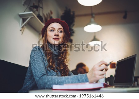 From below of glad female in red beret sitting at table with notebook and laptop while doing work task in cafeteria