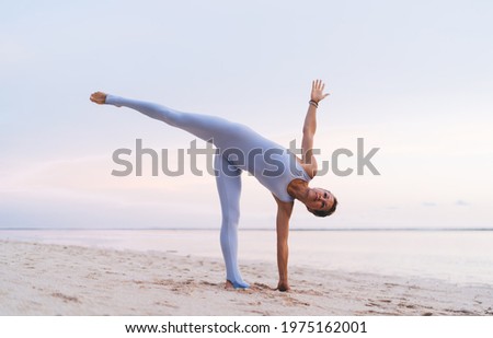 Side view from below of flexible barefoot female in sportswear doing yoga asana on sandy beach near water in summer