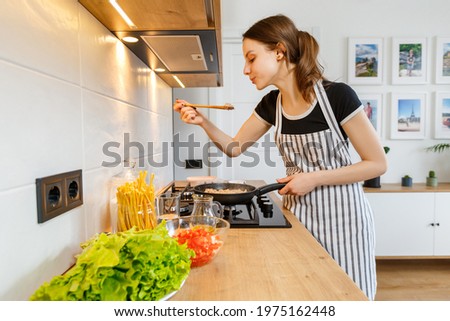Young woman in apron cooking healthy food at modern home kitchen. Preparing meal with frying pan on gas stove. Concept of domestic lifestyle, happy housewife leisure and culinary hobby. Royalty-Free Stock Photo #1975162448