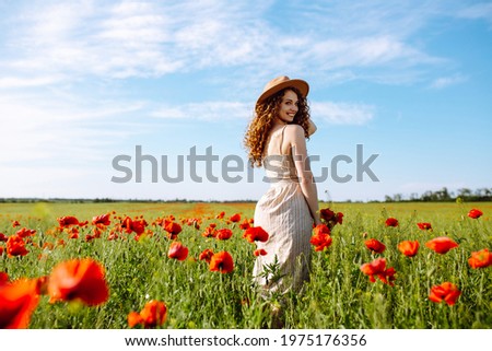 Beautiful girl posing in a poppy field. A woman with a hat stands in the middle of flowers. The young girl smiles. Happiness concept. Relaxing on summer poppy flowers meadow