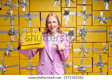 Charming woman representing new handbag in shop, pointing finger, posing at camera surrounded by gifts boxes. Hurry up while there are discounts