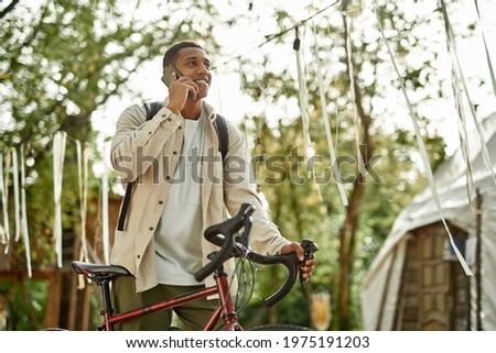 Young african american man talking on phone while standing next to bicycle in blooming nature with tent on background. Work in nature concept