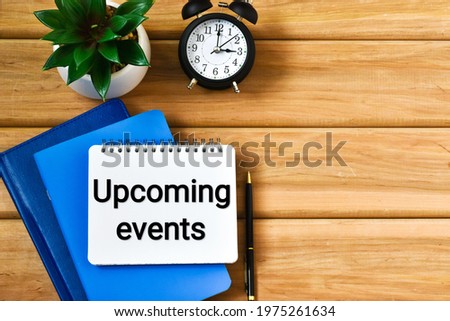 UPCOMING EVENTS text written in a notepad near a alarm clock, green plant, pen and a notebook on a wooden background. Business concept. Flat lay.