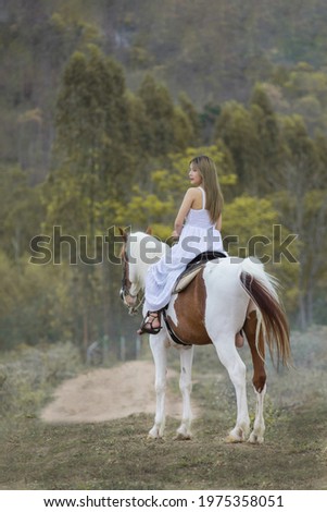 Asian woman in a white dress is riding a brown white horse. On the Countryside Behind the mountains