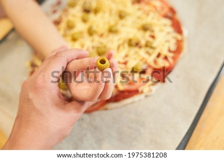 A mother holds a child's hand with an olive on the background of a healthy gluten-free homemade pizza, which is prepared for baking. Cooking together.