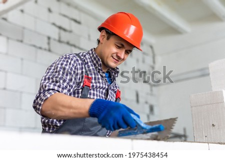 The process of bricklaying. Mason hands installing aerated concrete wall using special glue and tools necessary for brick works. Fresh mortar on aerocrete blocks. Brickwall layer with cement or glue. Royalty-Free Stock Photo #1975438454