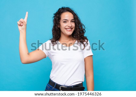 young pretty woman smiling cheerfully and happily, pointing upwards with one hand to copy space against blue background