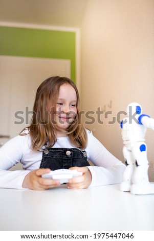 pretty young schoolgirl sitting on her desk in her room at home playing with small robot during corona time