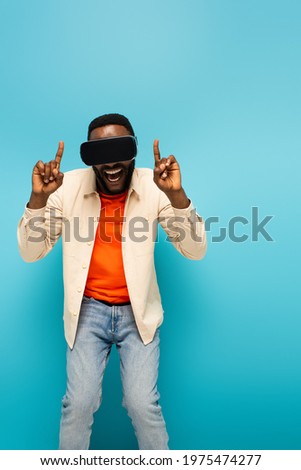 amazed african american man in vr headset pointing up with fingers on blue background