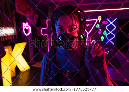 young asian woman in gas mask and wireless headphones looking at camera through metallic fence