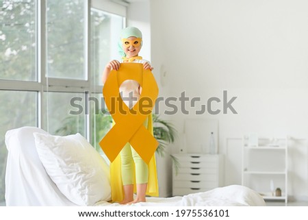 Little girl with golden ribbon wearing superhero costume in clinic. Childhood cancer awareness concept Royalty-Free Stock Photo #1975536101