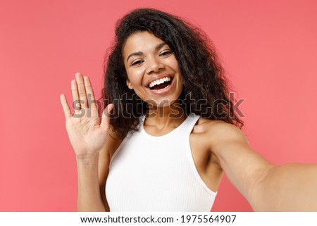 Close up young smiling friendly african american woman 20s in casual white tank shirt do selfie shot on mobile phone talk by video call waving hand greeting isolated on pink background studio portrait