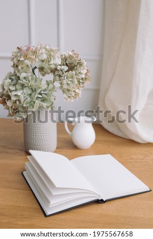Minimalist desk scene with modern ceramic vase with dry hydrangea, jug and journal open at  blank page.