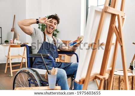 Young hispanic man sitting on wheelchair painting at art studio doing peace symbol with fingers over face, smiling cheerful showing victory 