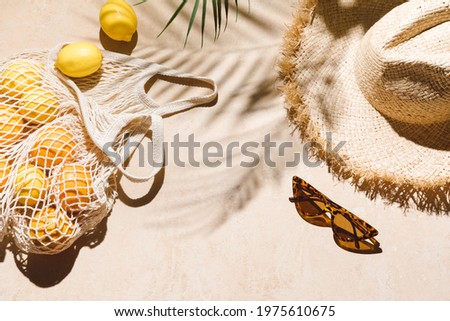 Summer flat lay on beige background. Straw hat, sunglasses, lemon fruits in eco friendly mesh shopping bag. Trendy palm shadow and sunlight, sun. Minimal summer travel fashion composition. Royalty-Free Stock Photo #1975610675