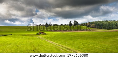 Landscape in the Hunsrueck, Germany. 
Field with green grass in front of trees. Landscape for banner. 
In the foreground grass with hills and storm clouds. 