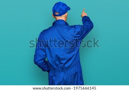 Bald man with beard wearing builder jumpsuit uniform posing backwards pointing ahead with finger hand 