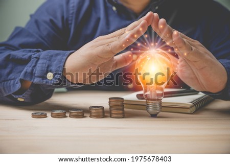 Concept accounting, saving money for business. Businessman Protect at a lightbulb with coins stacking and notebooks on a wooden table with. Idea saving energy and investment business into the future.