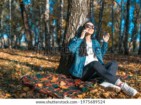 A brunette emotionally communicates on a mobile phone, a surprised woman in sunglasses is resting in a park near a tree in autumn. The concept of communication on a smartphone in nature. copy space
