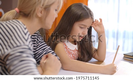 Child daughter 6-7 years old is studying with her young mother teacher. School kid girl teaching adult parent helping mom with studying homework sitting at desk at home. Children education concept.