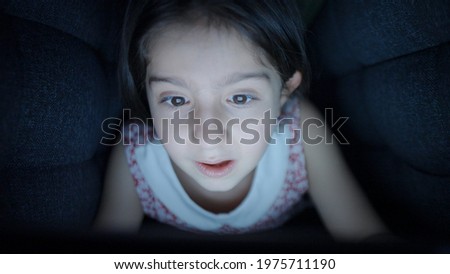 Beautiful little girl spends time with tablet computer or mobile phone in the dark under the duvet. Internet or online game addicted concept.