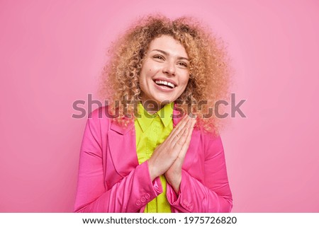 Sincere positive curly haired young woman keeps palms pressed together laughs happily expresses joy feels very glad as being promoted at work dressed elegantly isolated over pink background.