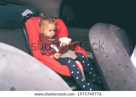 Cute caucasian little child sitting at safety car seat for children behind driver seat, fastened by safety belts, watching cartoons and videos at digital device, evening time, holding bear toy.