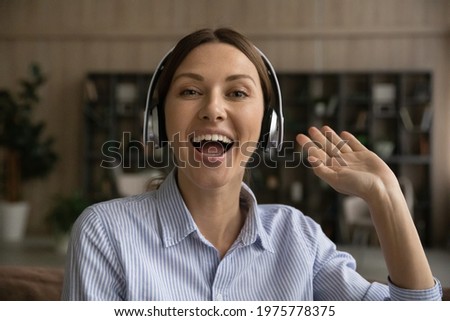 Hi everyone. Portrait of active teen female presenter in headphones look at camera greet gen z audience from screen. Positive young lady vlogger influencer welcome subscriber on personal video channel Royalty-Free Stock Photo #1975778375