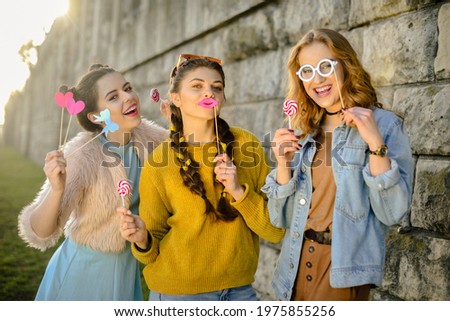 Hipster girls, colored dressed, with sunglasses and wrist watch. Grup of three girls with funny lips, glasses, stars, paper hearts on stick at the sunset having fun. Going crazy, smile at summer day. 