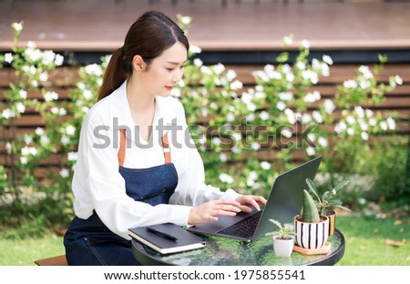 Young asian gardener working with laptop to record and note on book the growth of trees.Cute asian Greenhouse worker or owner small business selling small tree startup and sustainable concept.