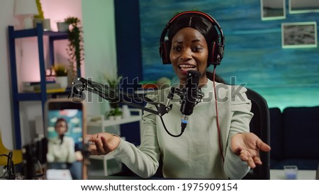 Focused shot on smartphone recording african blogger woman talking looking at camera during online podcast. Content creator streaming online broadcast, blogger discussing wearing headphones.