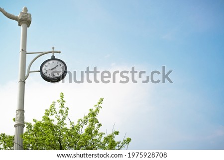 Clock hanging on a vintage pole, outdoors on a sunny day, blue clear sky background. Black dial showing the time outside. Copy space for text