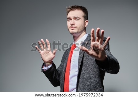 Serious man in a formal suit covers himself with his hands from the camera, showing a stop sign. Business concept. Studio portrait on a grey background. 