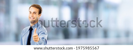 Smiling confident young businessman showing thumbs up like hand sign gesture, over blurred modern office interior background. Happy confident man gesturing. Copy space area. Success in business. 