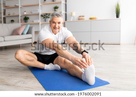 Sport at home during quarantine. Senior man doing stretch exercises for legs, working out and sitting on mat in living room interior and smiling to camera, free space Royalty-Free Stock Photo #1976002091