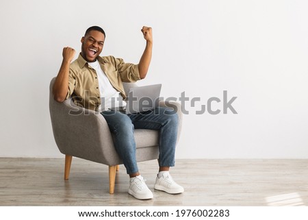 Great News. Emotional African American Guy With Laptop Computer Gesturing Yes Celebrating Success Sitting In Armchair Over Gray Wall Indoors. Looking At Camera. Luck, Joy Of Victory