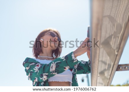 Outdoor portrait of woman in casual green shirt at sunny day stands on bridge looking on phone screen take selfie make video call wireless bluetooth headphones in ears