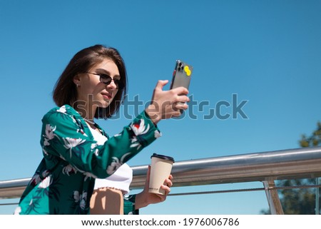 Outdoor portrait of woman in casual green shirt and jeans at sunny day walks on bridge with cup of coffee looking on phone screen