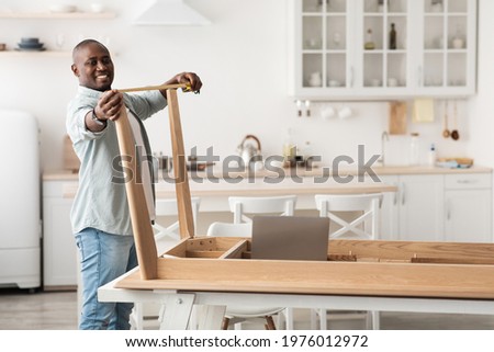 Happy black man measuring the width between legs of wooden table, watching online instructions on laptop computer while assembling furniture after relocation in kitchen, free space Royalty-Free Stock Photo #1976012972